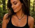 A woman wearing a black cord necklace with three silver-toned claw-shaped pendants, photographed outdoors in a sunlit forest setting.