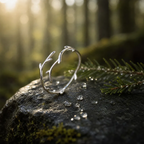A silver ring designed to look like small branches or antlers, resting on a mossy rock with pine needles and water droplets in a sunlit forest setting.