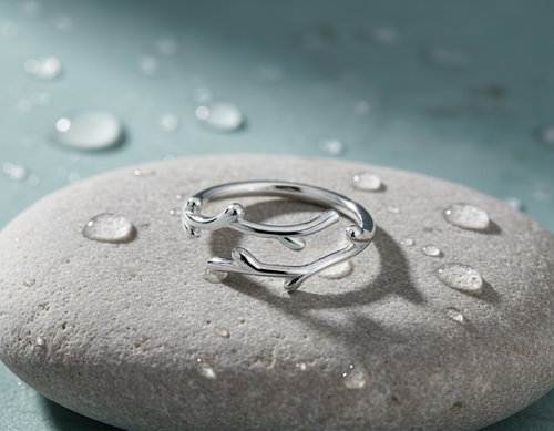 A silver ring with a branch-like, organic open design, resting on a smooth gray stone covered in water droplets against a soft teal-blue background.