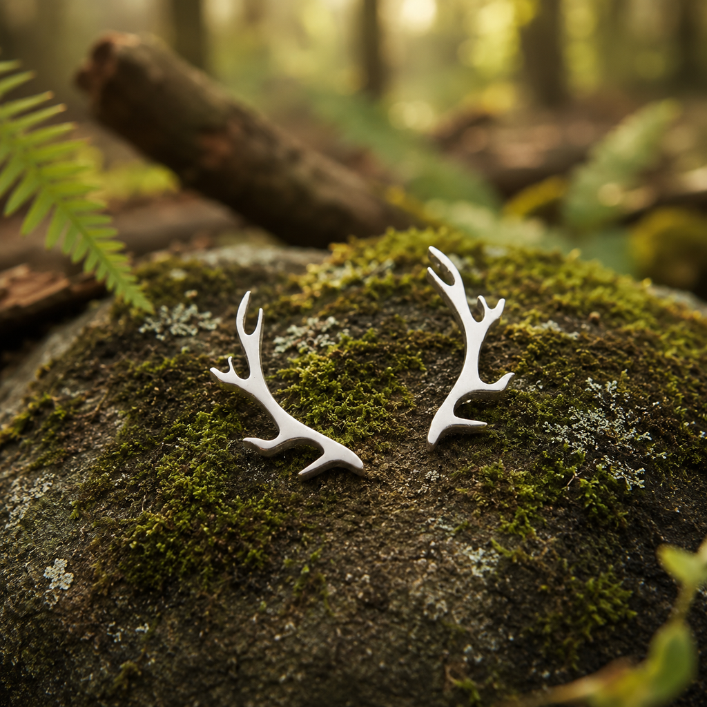 A pair of silver deer antler stud earrings resting on a mossy forest rock with a blurred woodland background.