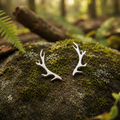 A pair of silver deer antler stud earrings resting on a mossy forest rock with a blurred woodland background.