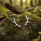 A pair of silver deer antler stud earrings resting on a mossy forest rock with a blurred woodland background.