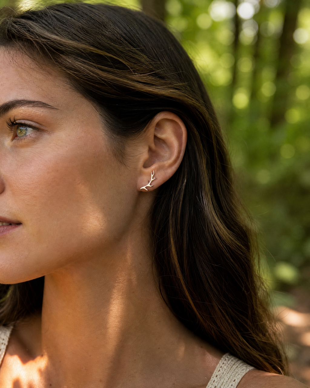 A close-up side profile of a woman with long brown hair wearing a delicate silver leaf-shaped ear cuff, set against a blurred green forest background.