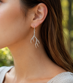 A close-up of a woman's ear wearing a delicate silver antler dangle earring, with a soft-focus outdoor background of green foliage.