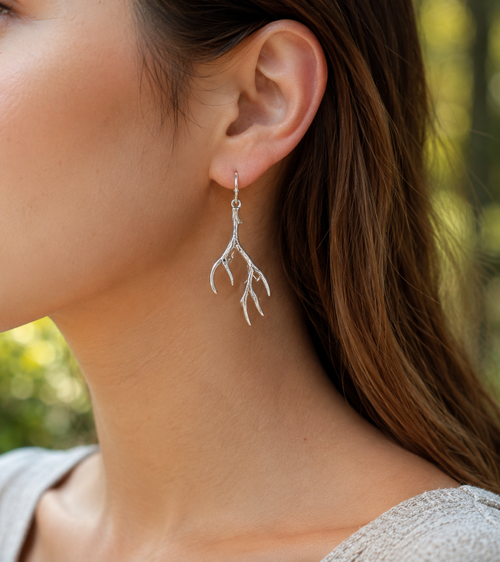 A close-up of a woman's ear wearing a delicate silver antler dangle earring, with a soft-focus outdoor background of green foliage.