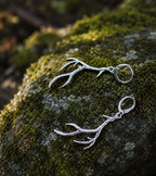 A pair of silver-toned antler drop earrings resting on a moss-covered rock outdoors.