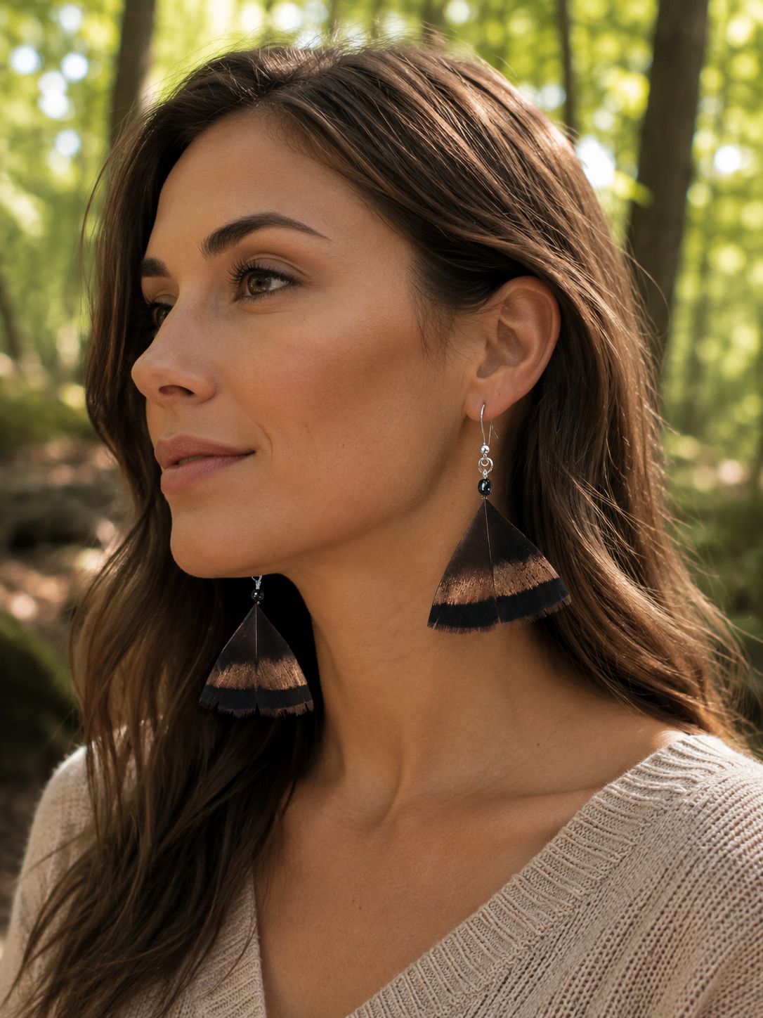 A woman in a beige knit top wearing large dark brown and copper feathered dangle earrings with silver hooks, photographed outdoors in a sunlit forest setting.
