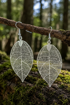 A pair of silver leaf-shaped dangle earrings with intricate openwork vein detailing, hanging from a mossy tree branch in a forest setting.