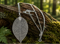 A silver leaf-shaped pendant necklace with an intricate openwork design, displayed on a mossy log in a forest setting.