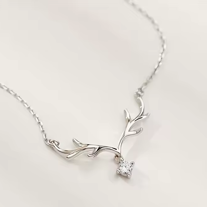 A delicate silver necklace featuring a branch-shaped pendant with a single small clear gemstone, displayed on a soft, light beige background.
