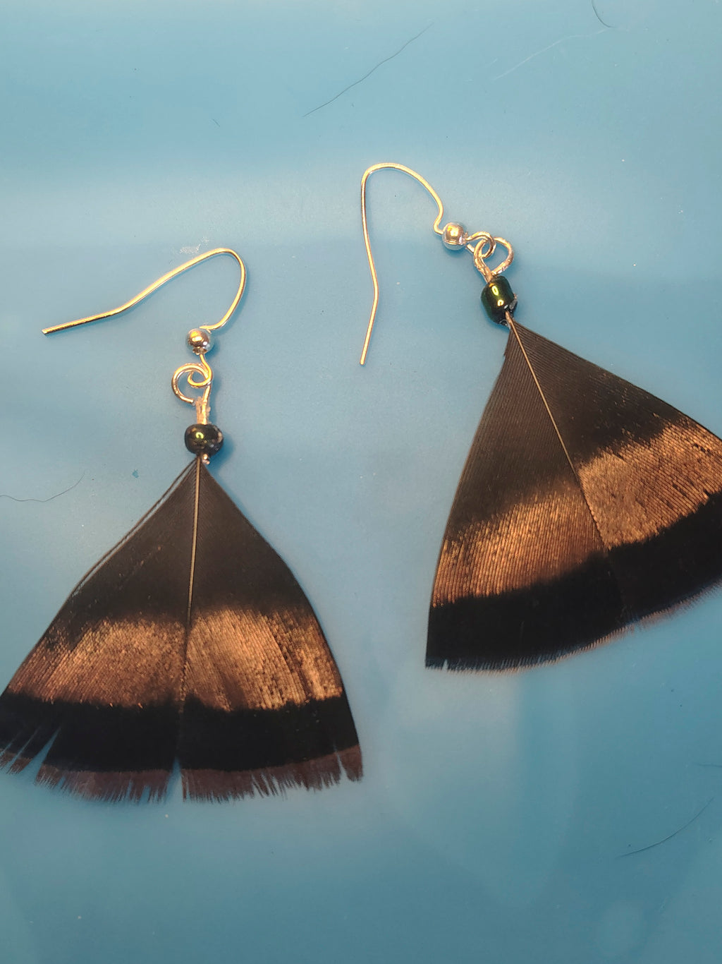 A pair of dangle earrings featuring large brown, copper and black turkey feather fan shapes suspended from gold fishhook wires with small black beads, displayed on a smooth light blue surface.