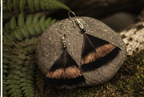 A pair of dangling fan-shaped earrings made from dark brown  feathers with metallic copper-brown bands, hanging from silver hooks and displayed on a round gray stone with green ferns in the background.
